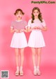 Two young women standing next to each other on a pink background.